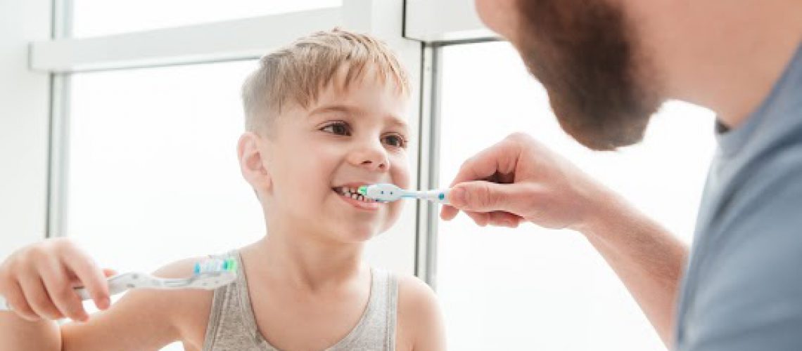 Photo of father and son smiling while brushing teeth in bathroom. Look at each other.